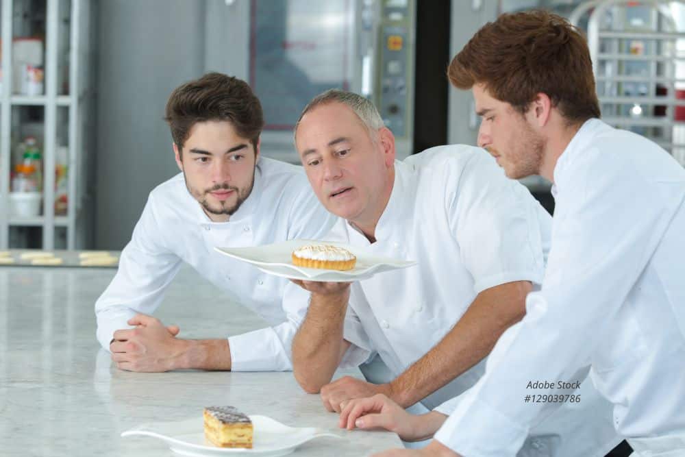 Abbildung: Zwei Auszubildende mit Ihrem Ausbilder betrachten ein Törtchen in der Bäckerei.